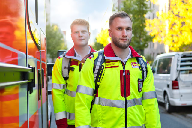 Bild zeigt zwei Mitarbeiter des ASB seitlich neben dem RTW, in Dienstkleidung stehend. 